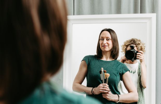 Makeupartistin Diana Schulte mit Pinseln in der Hand steht vor einem Spiegel. Man sieht sie und die Fotografin Rebecca Peetz im Spiegel, während sie beide fotografiert.